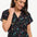 A woman smiling and wearing a black blouse with a colorful floral and star print, featuring a round grandad collar and short cap sleeves.