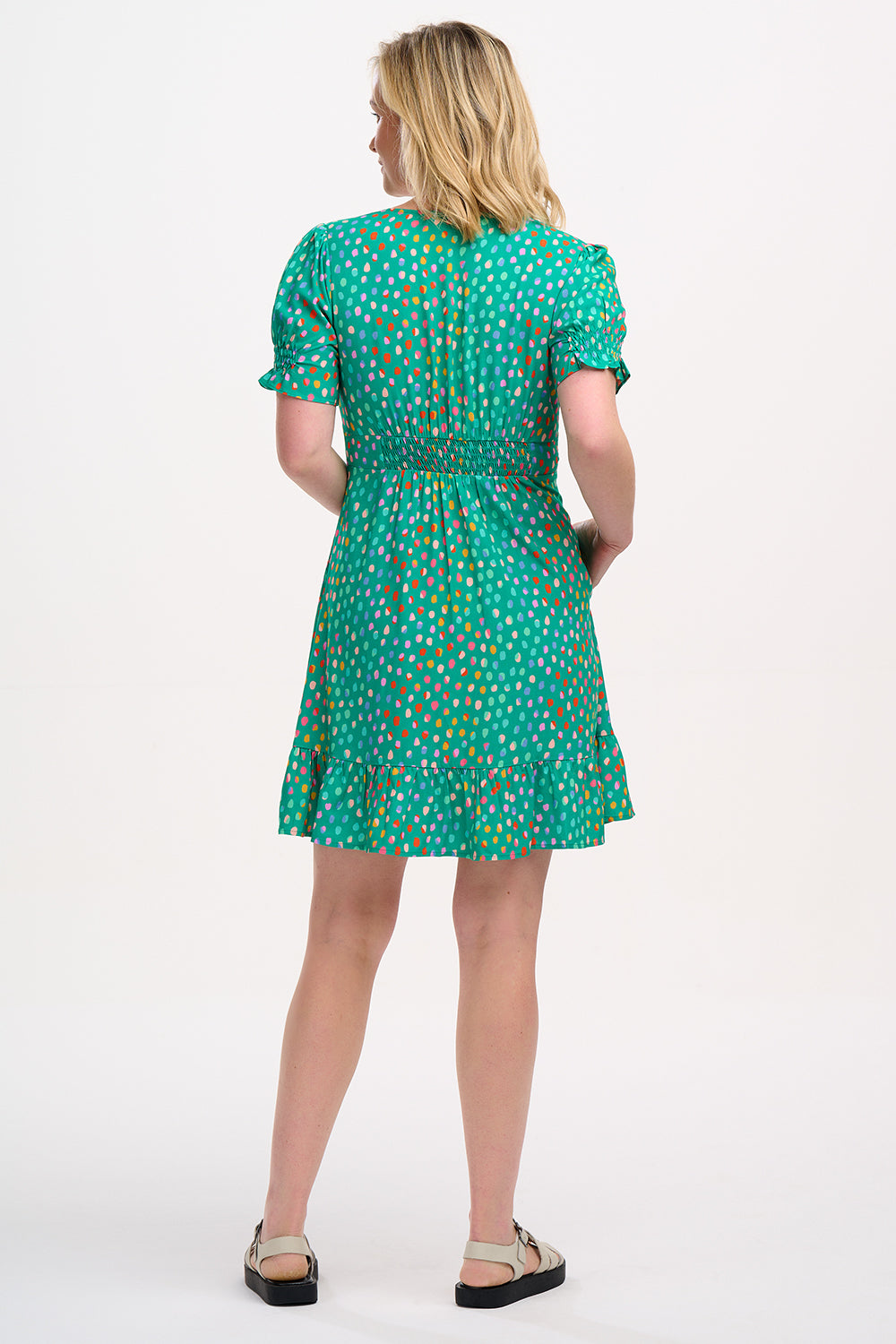 A woman wearing a green mini dress with a pattern of rainbow pebbles, featuring a V-neckline, puff sleeves, and a frill hem.