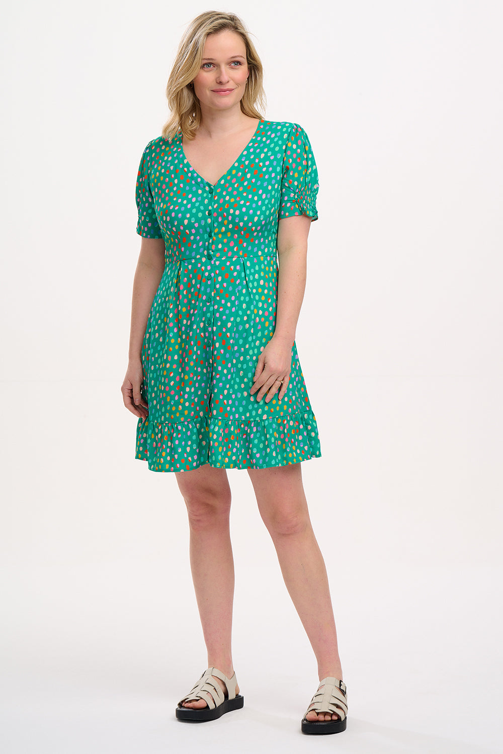 A woman wearing a green mini dress with a pattern of rainbow pebbles, featuring a V-neckline, puff sleeves, and a frill hem.