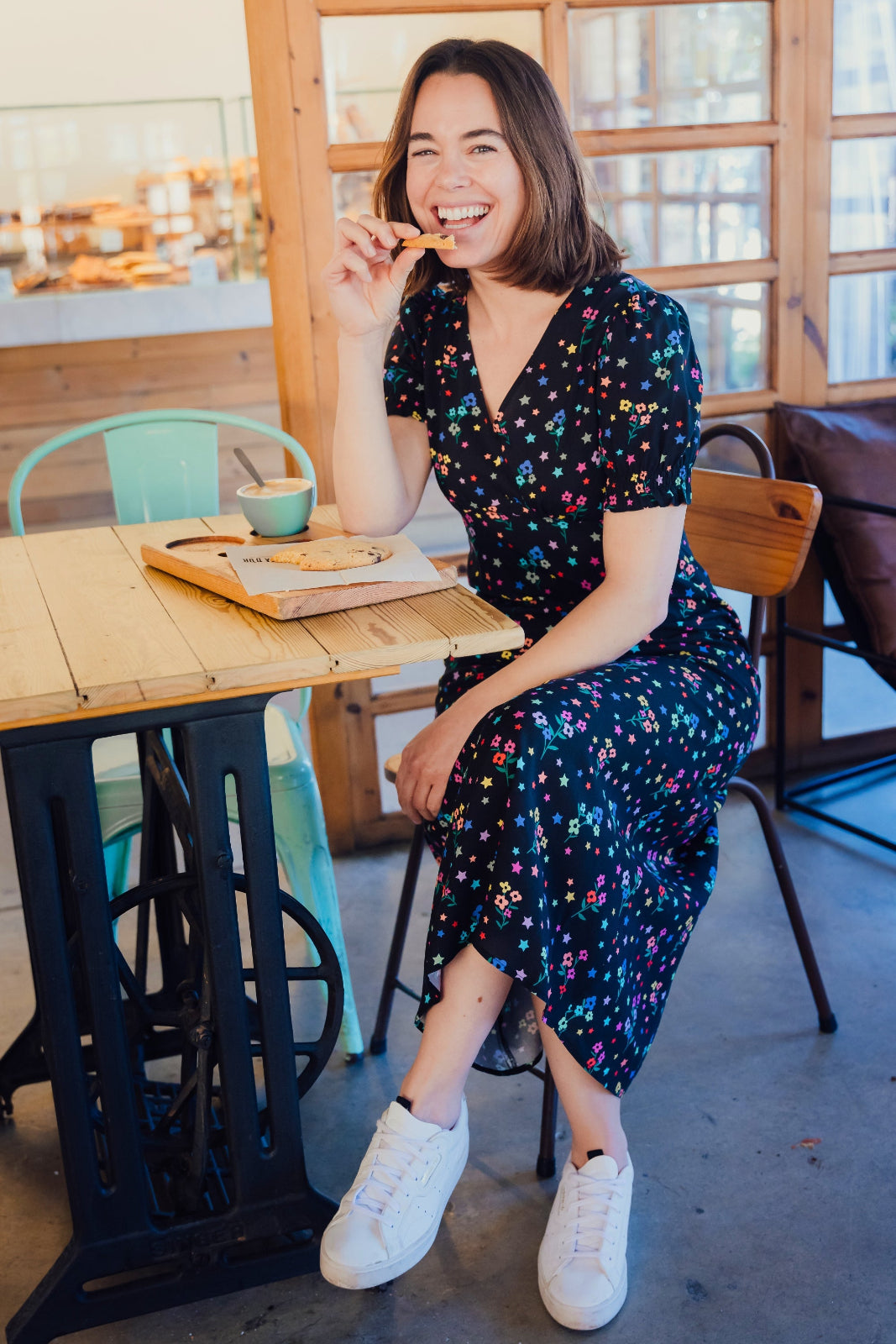 A woman sitting at a cafe wearing a black midi dress with a rainbow sprigs print, featuring a V-neck and short-shirred cuffs.