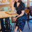 A woman sitting at a cafe wearing a black midi dress with a rainbow sprigs print, featuring a V-neck and short-shirred cuffs.