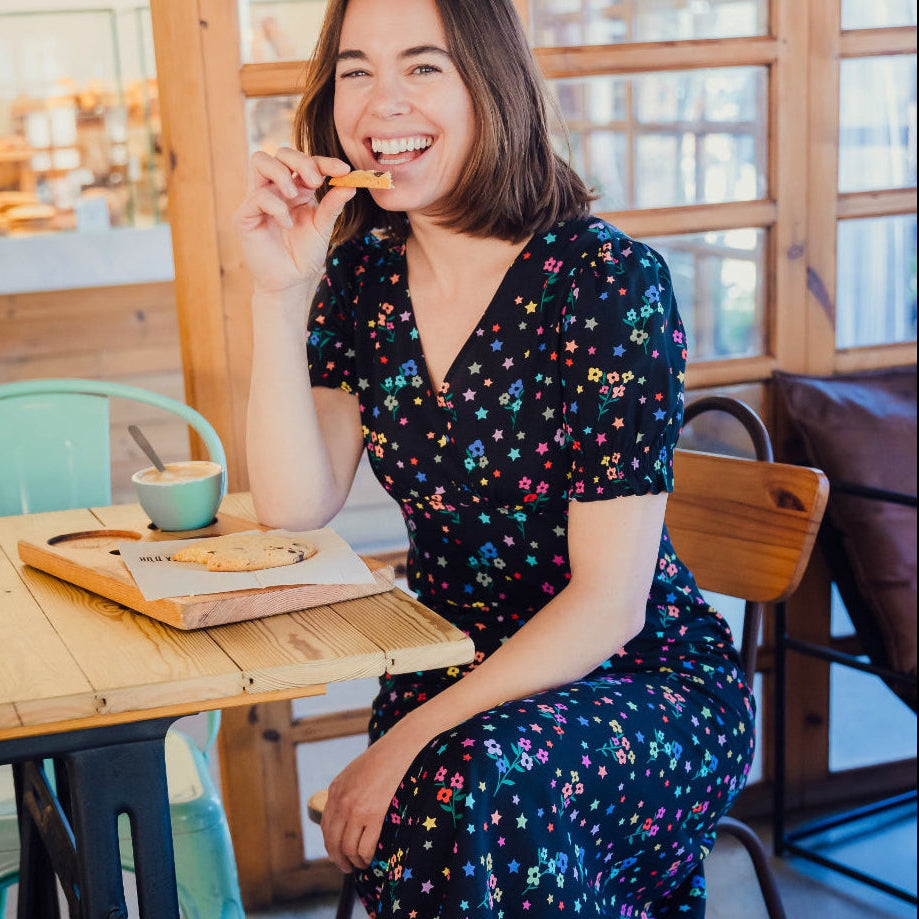 A woman sitting at a cafe wearing a black midi dress with a rainbow sprigs print, featuring a V-neck and short-shirred cuffs.