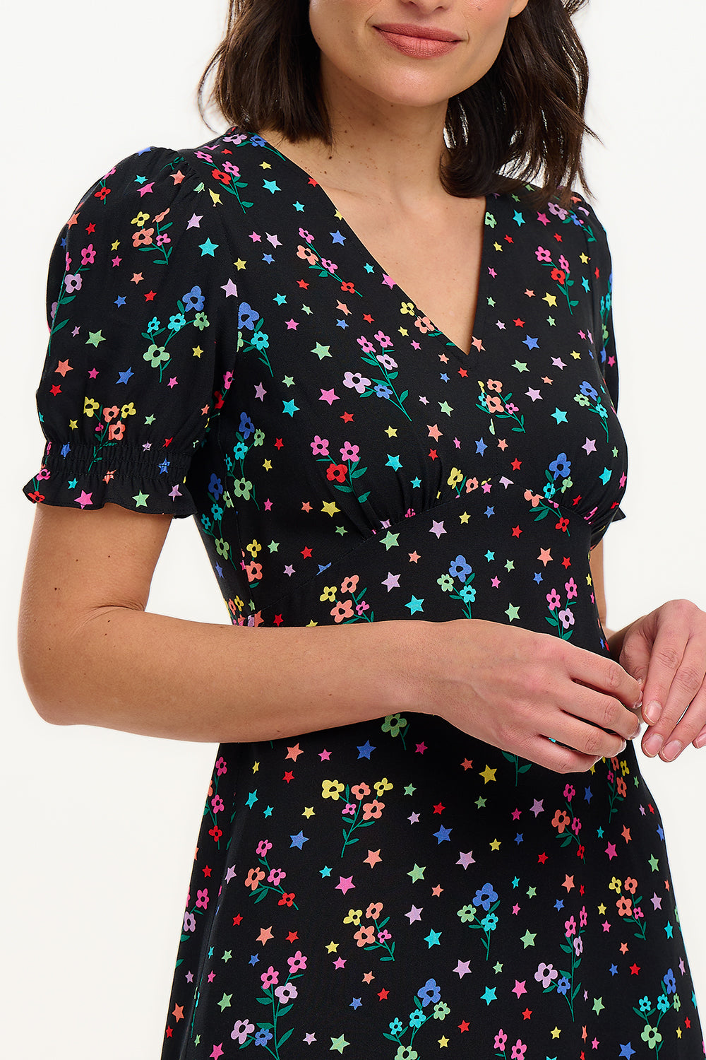 A model wearing a mid-length black dress with a V-neck and short-shirred sleeves, adorned with a multicolor floral and star print.