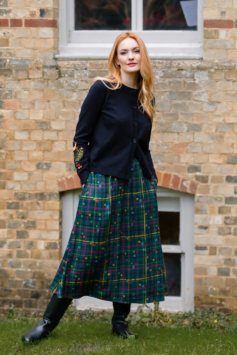 A woman stands outdoors in front of a brick building, wearing a black long-sleeve top, a green plaid midi skirt with yellow and red lines, and black boots.