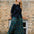 A woman stands outdoors in front of a brick building, wearing a black long-sleeve top, a green plaid midi skirt with yellow and red lines, and black boots.