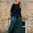 A woman stands outdoors in front of a brick building, wearing a black long-sleeve top, a green plaid midi skirt with yellow and red lines, and black boots.