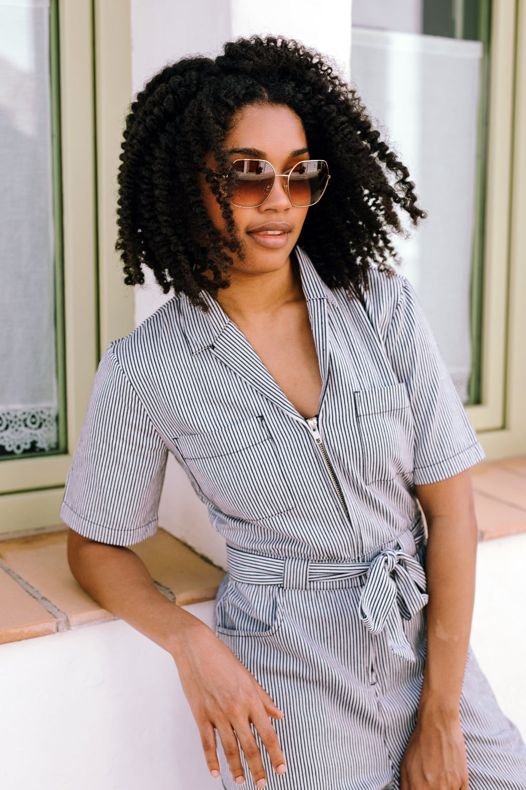 Woman wearing a striped shirt jumpsuit and sunglasses sitting on a bench.