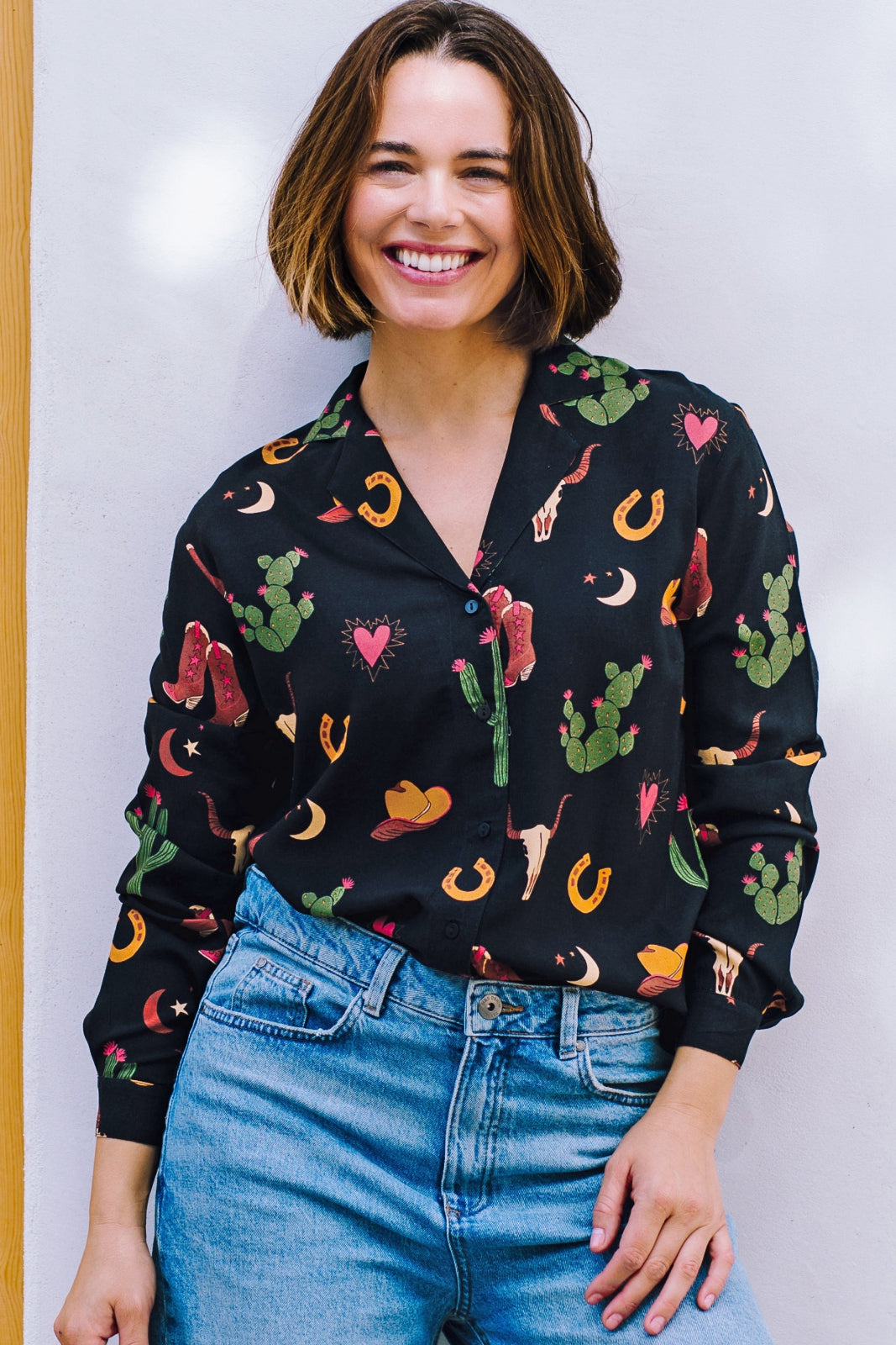 A woman wearing a black long-sleeved shirt with a cowgirl print of cactus, hearts, cowboy boots, and horseshoes, paired with blue jeans.