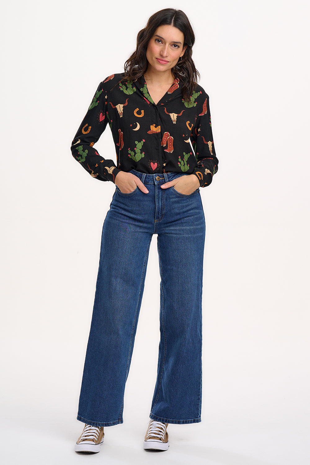 A woman wearing a black long-sleeved shirt with a cowgirl print of cactus, hearts, cowboy boots, and horseshoes, paired with blue jeans.