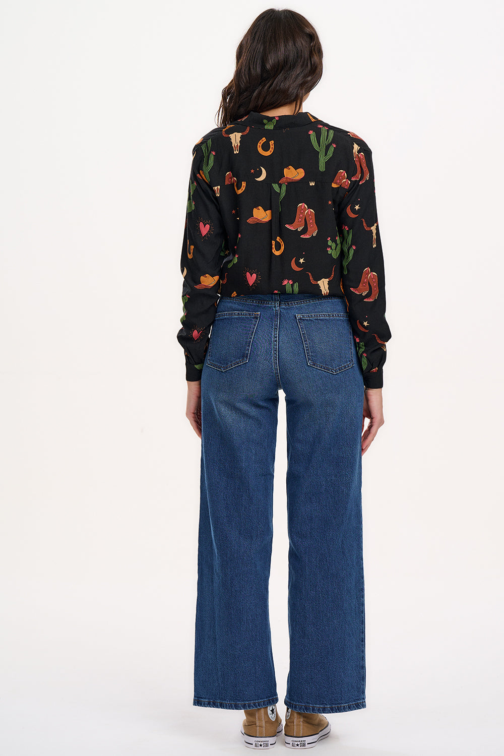 A woman wearing a black long-sleeved shirt with a cowgirl print of cactus, hearts, cowboy boots, and horseshoes, paired with blue jeans.