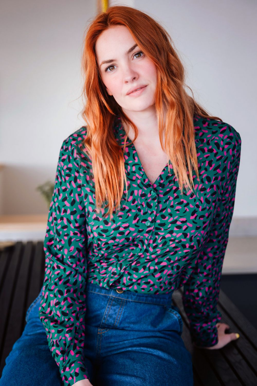 Woman with red hair wearing a patterned blouse sitting on a bench.
