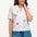 A model wearing an off-white shirt with a colorful fruit and cocktail themed pattern, paired with blue denim shorts.