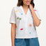 A model wearing an off-white shirt with a colorful fruit and cocktail themed pattern, paired with blue denim shorts.