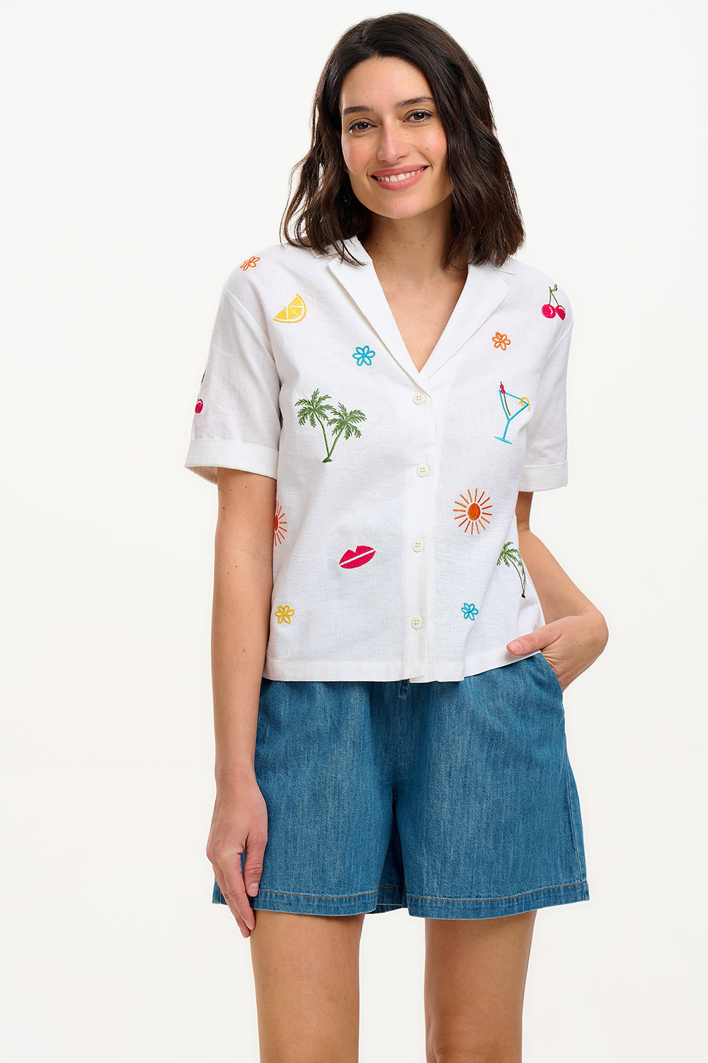 A model wearing an off-white shirt with a colorful fruit and cocktail themed pattern, paired with blue denim shorts.