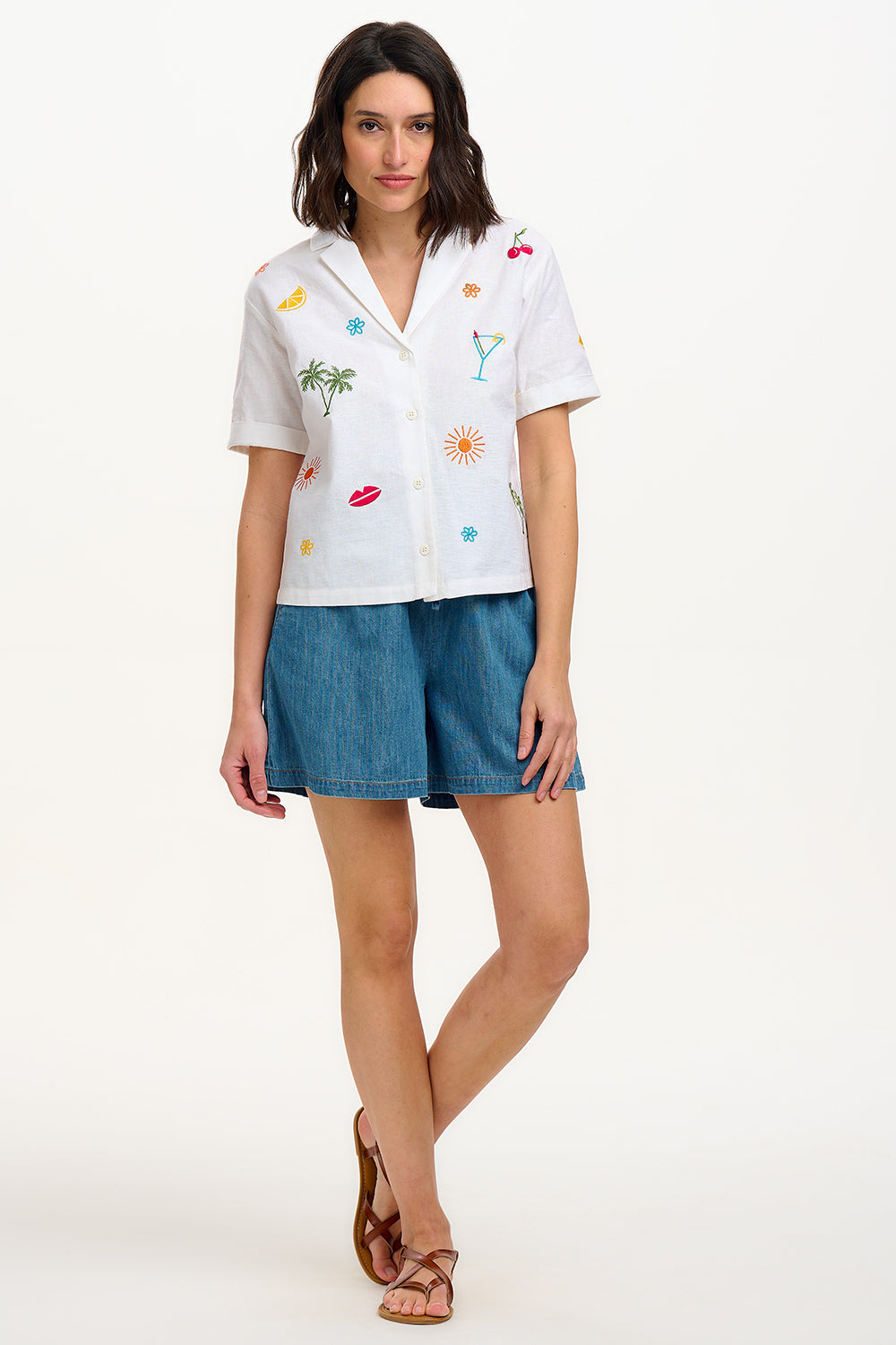 A model wearing an off-white shirt with a colorful fruit and cocktail themed pattern, paired with blue denim shorts.