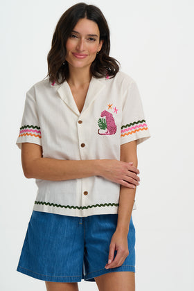 Woman wearing a white embroidered shirt with colorful accents and blue shorts on a white background