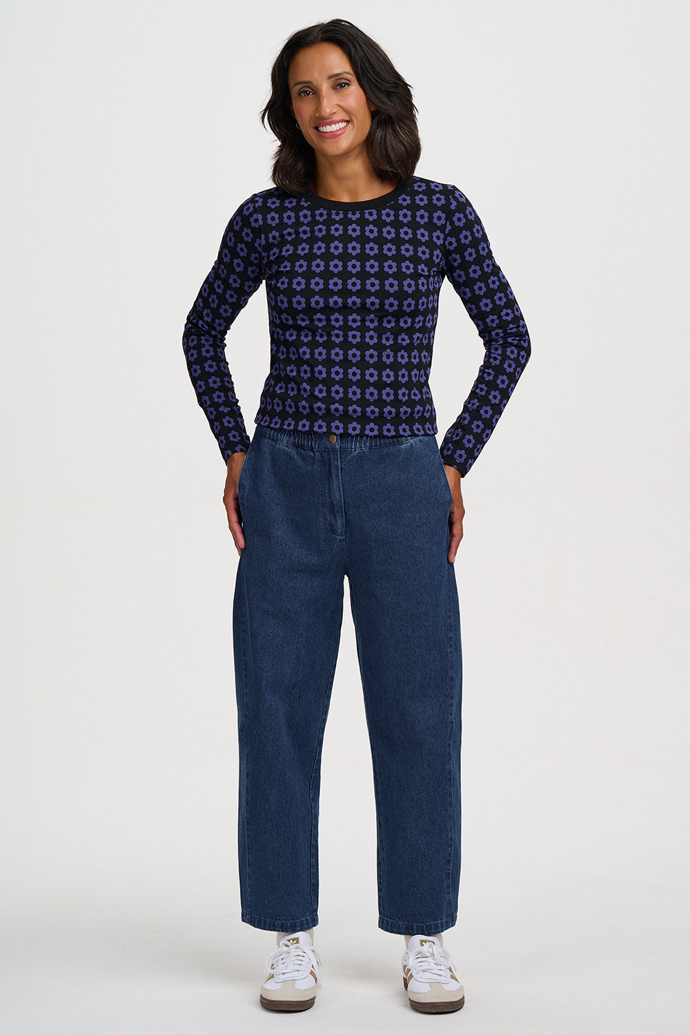 A woman modeling a navy blue long-sleeve top with a small white geometric pattern, paired with dark blue jeans and white sneakers, against a light gray studio background.