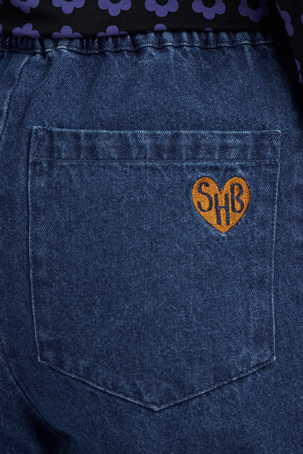 Close-up of the back pocket of dark blue denim jeans, featuring a small orange embroidered heart patch with the letters 'SHB' in black.