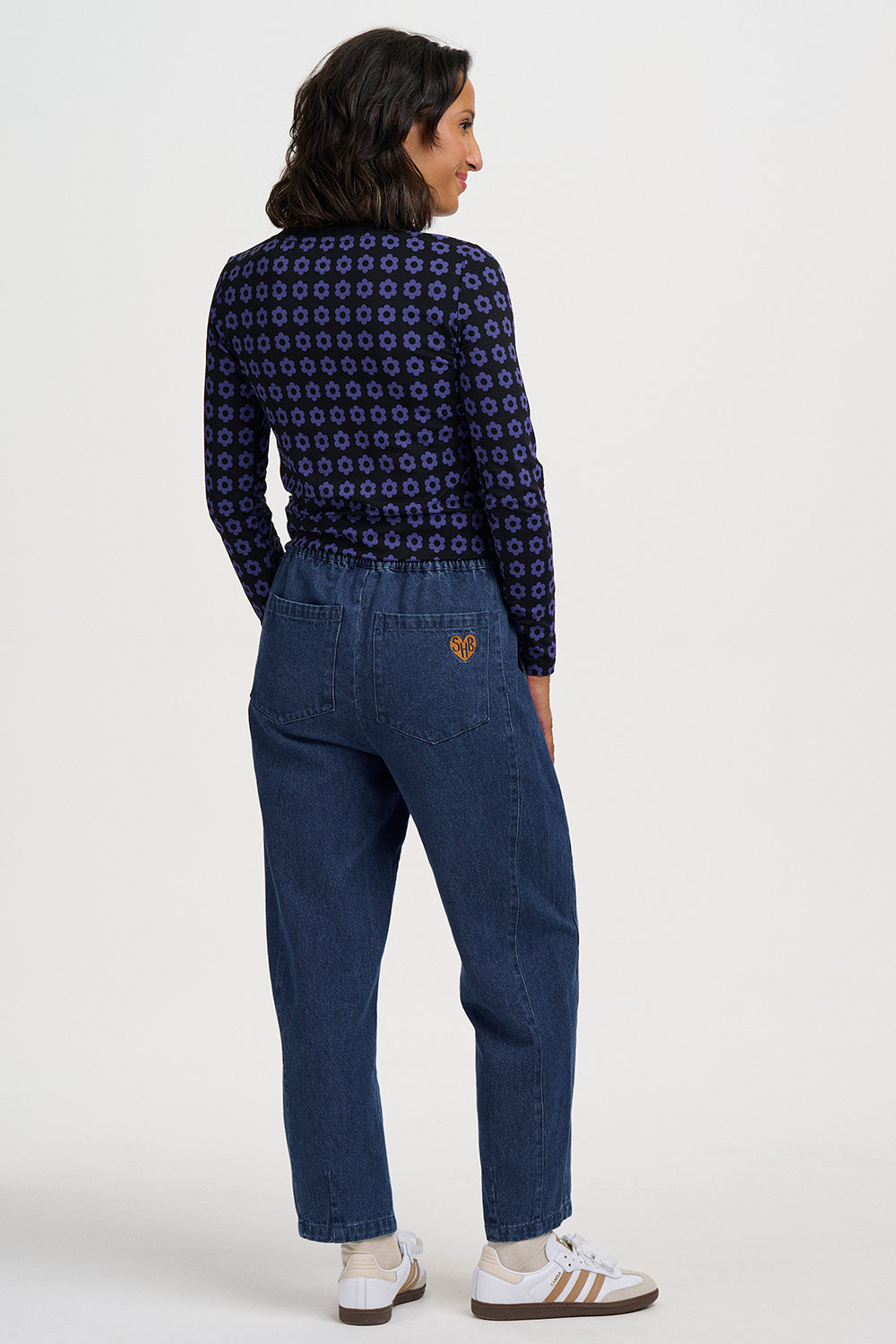 A model is shown from the back wearing dark blue jeans with a small gold embroidered logo on the back pocket, paired with a navy patterned long-sleeve shirt and white sneakers.