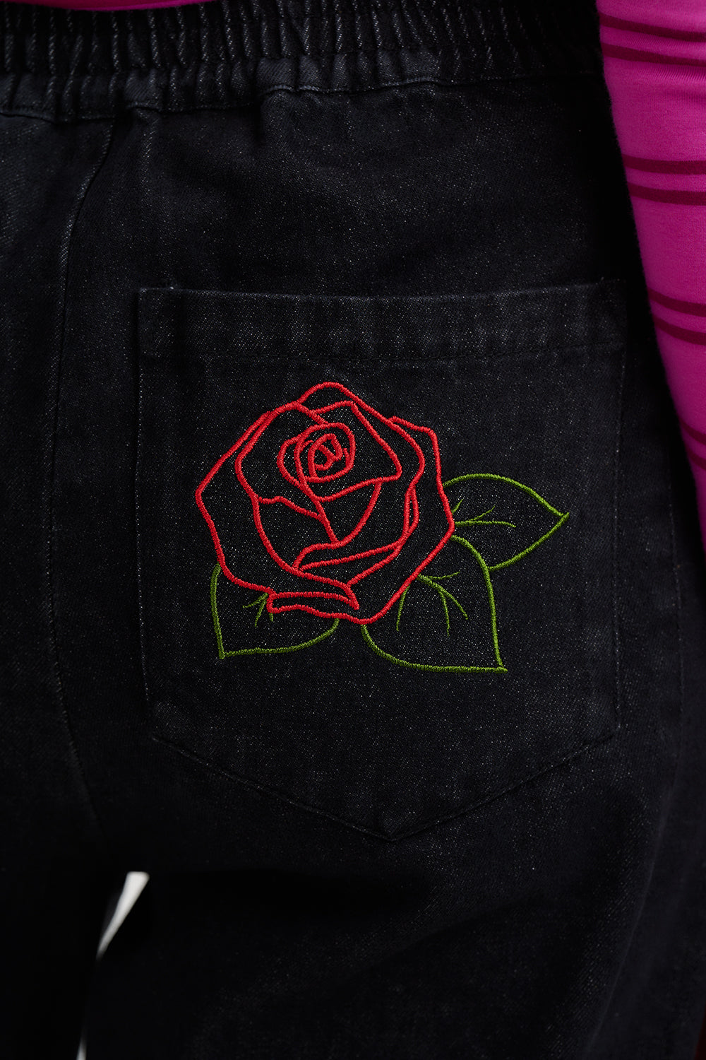 Black pants with a red rose embroidery on the pocket, against a pink background.