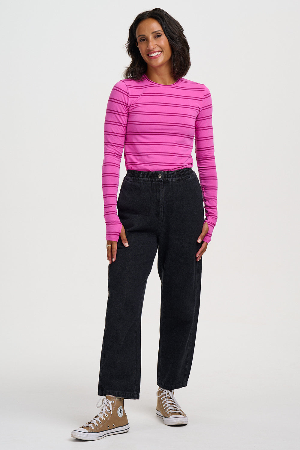 A woman modeling a bright pink long-sleeve knit top with thin dark horizontal stripes, paired with black jeans and tan sneakers, against a plain light gray studio background.