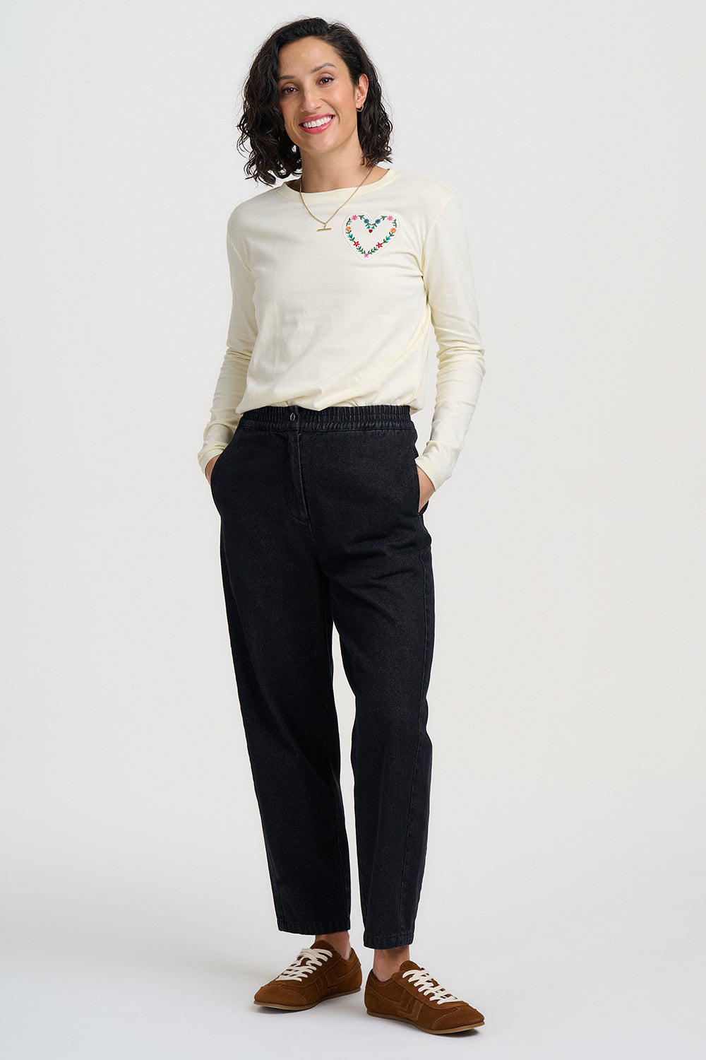 Woman wearing a cream sweater with a heart design and black pants on a white background