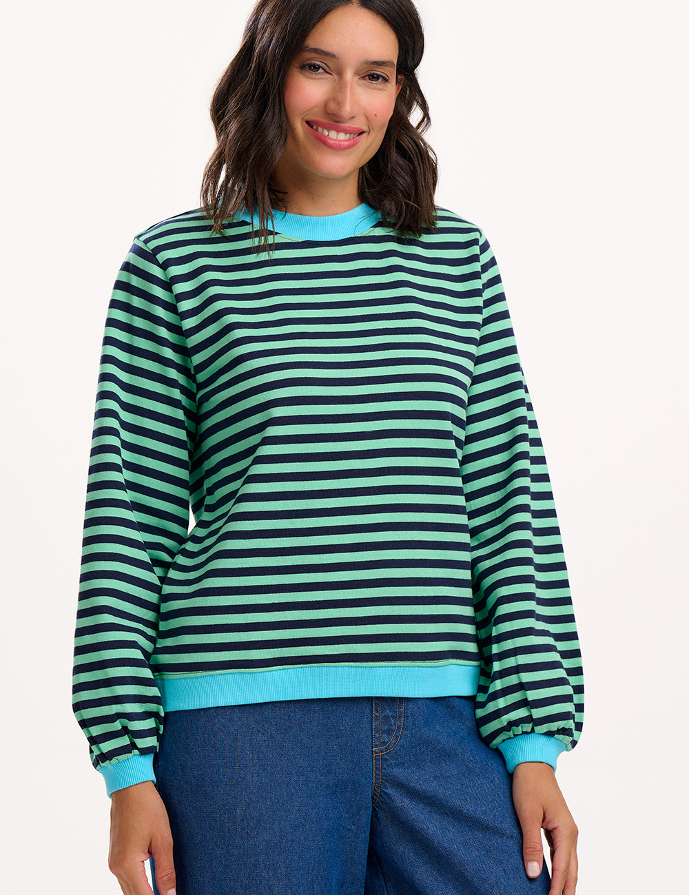 Sugarhill Brighton model wearing a green and blue striped sweater with blue jeans on a white background