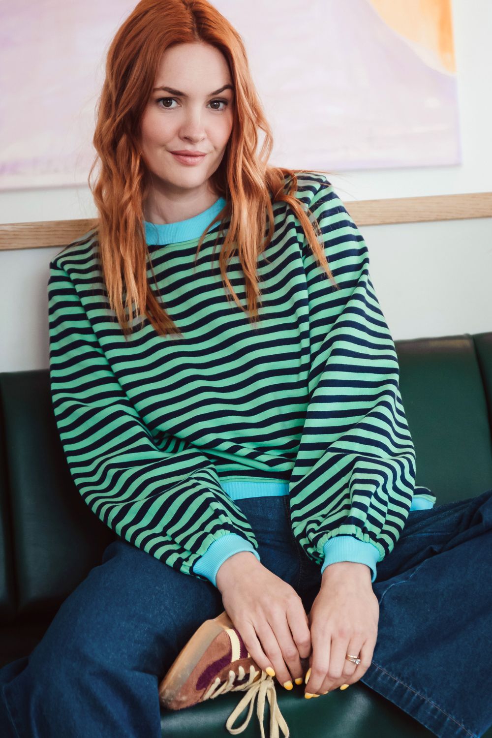 Sugarhill Brighton model wearing green and blue striped sweatshirt on a sofa. 
