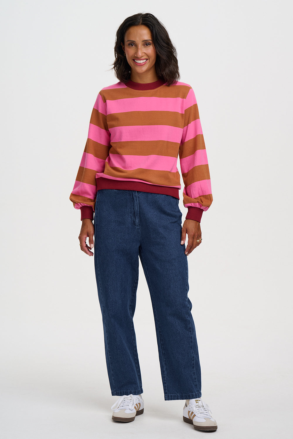 A woman modeling a pink and brown horizontally striped long-sleeve sweatshirt with dark blue jeans and white sneakers, standing against a light gray studio background.