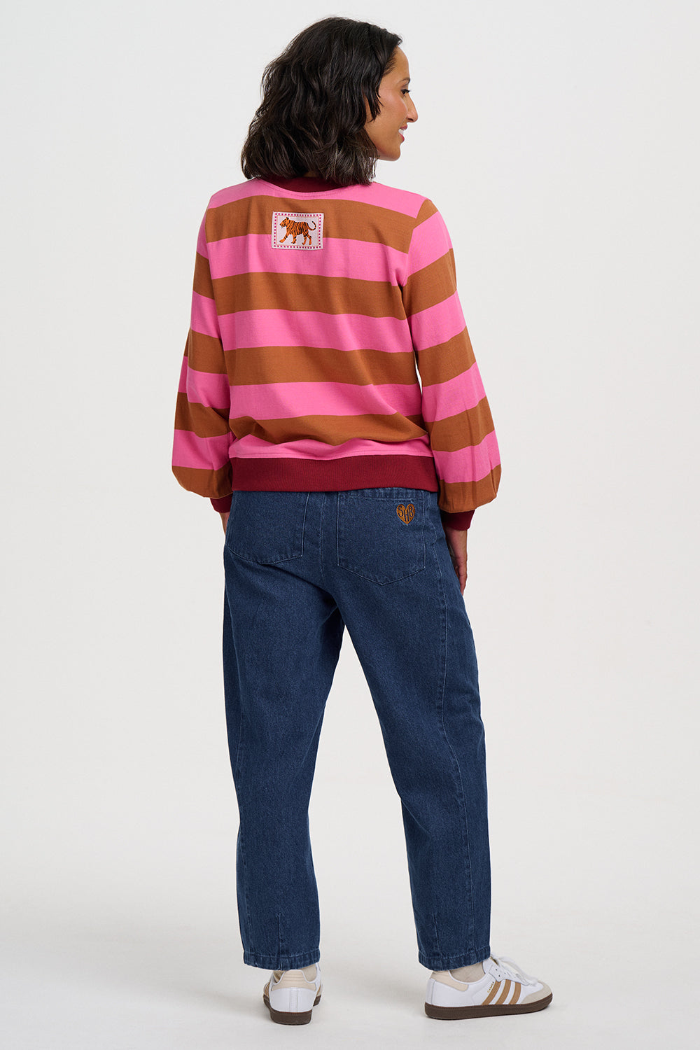 A woman shown from the back wearing a pink and brown horizontally striped sweatshirt with a small horse patch on the back, blue jeans, and white sneakers, against a plain light gray studio background.