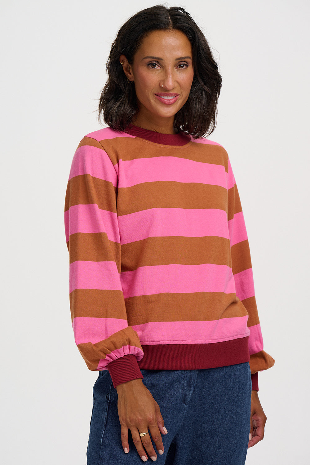 A woman wearing a pink and brown horizontally striped long-sleeve sweatshirt with dark red ribbed trim, paired with blue jeans, against a light gray studio background.