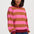 A woman wearing a pink and brown horizontally striped long-sleeve sweatshirt with dark red ribbed trim, paired with blue jeans, against a light gray studio background.
