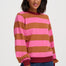 A woman wearing a pink and brown horizontally striped long-sleeve sweatshirt with dark red ribbed trim, paired with blue jeans, against a light gray studio background.