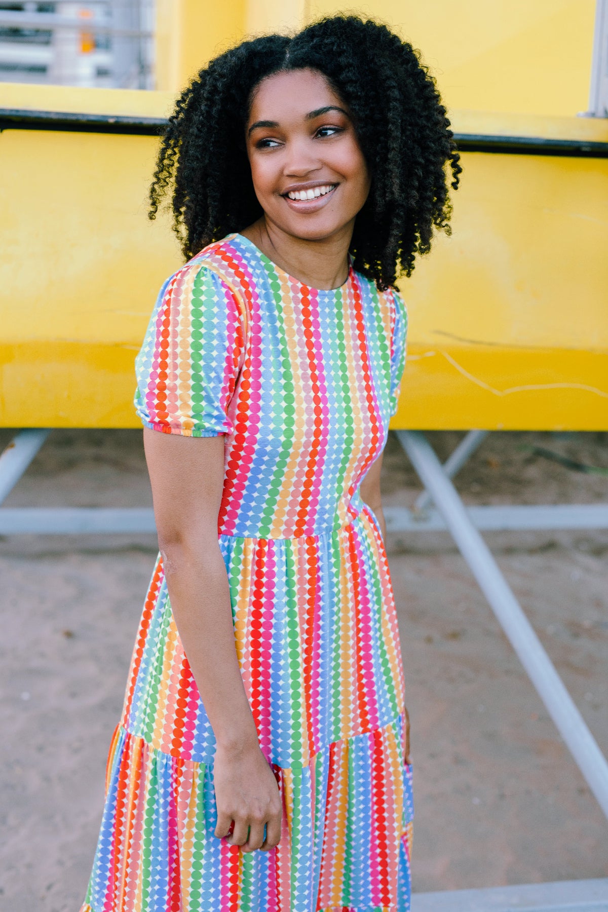 A woman smiling and wearing a multicolored tiered midi jersey dress with a round neckline and short puff sleeves.