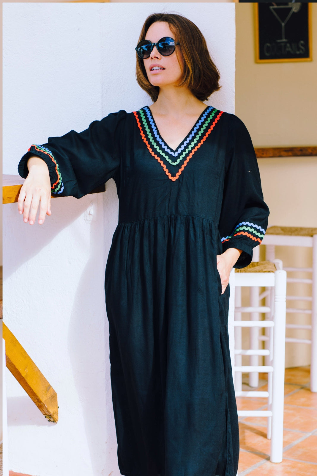 A model wearing a midi length black smock dress with rainbow Ric Rac detailing around the neckline and sleeve cuffs.