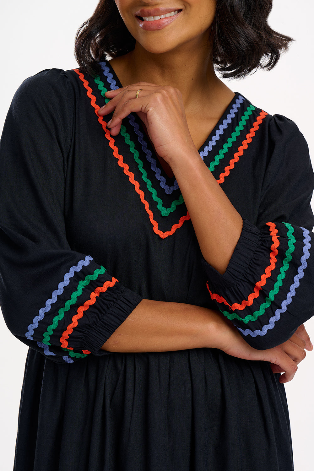 A model wearing a midi length black smock dress with rainbow Ric Rac detailing around the neckline and sleeve cuffs.