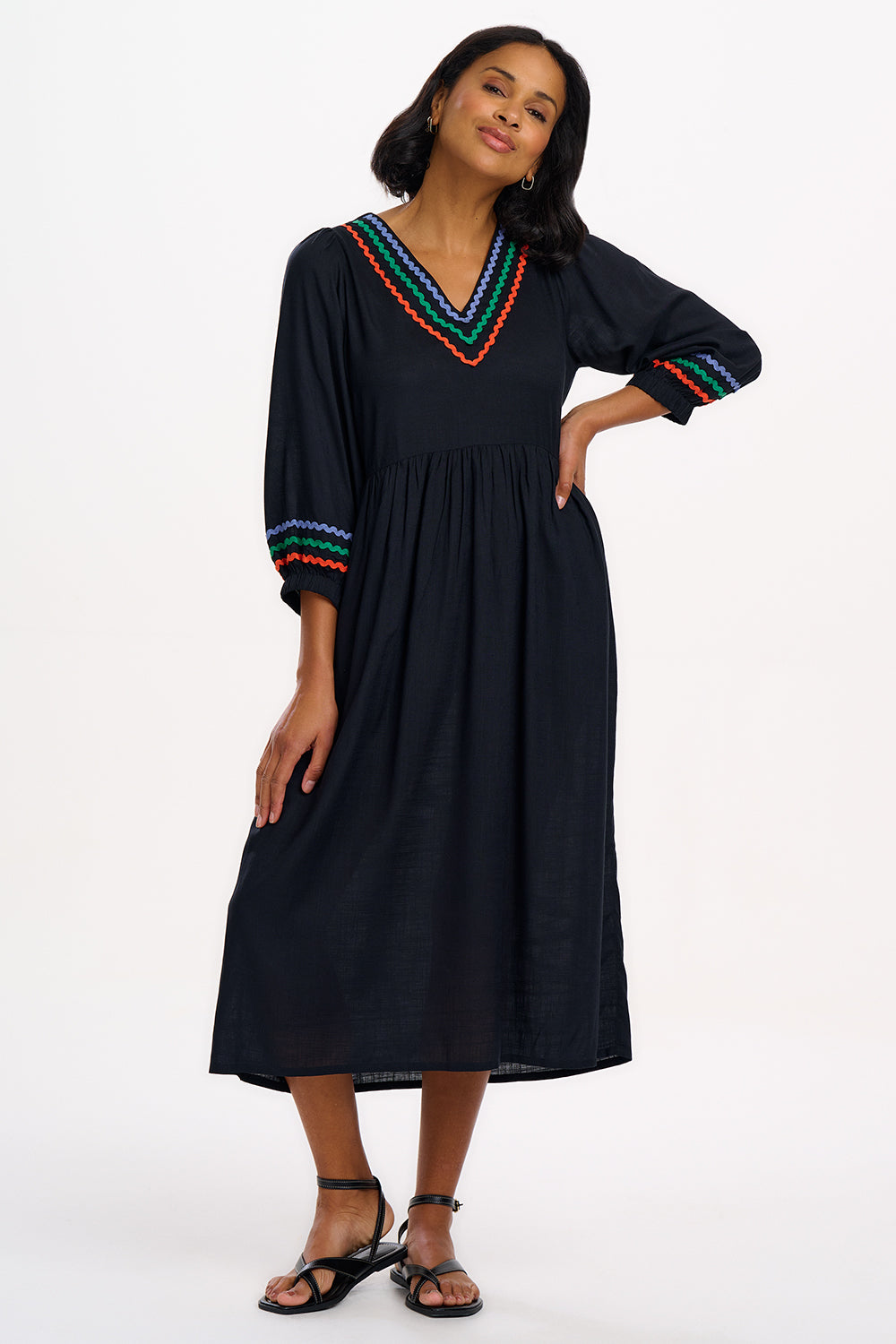 A model wearing a midi length black smock dress with rainbow Ric Rac detailing around the neckline and sleeve cuffs.