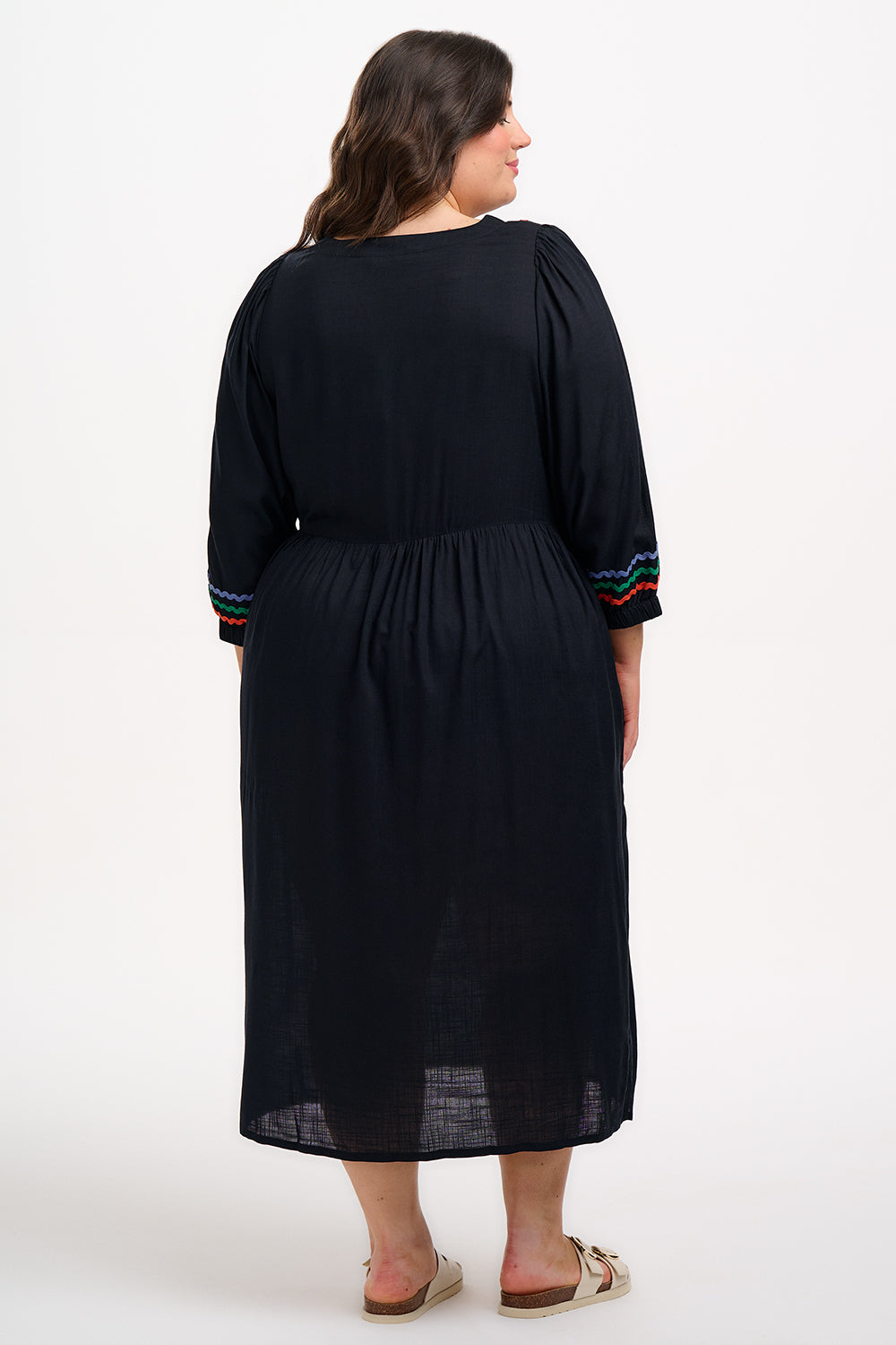 A model wearing a midi length black smock dress with rainbow Ric Rac detailing around the neckline and sleeve cuffs.