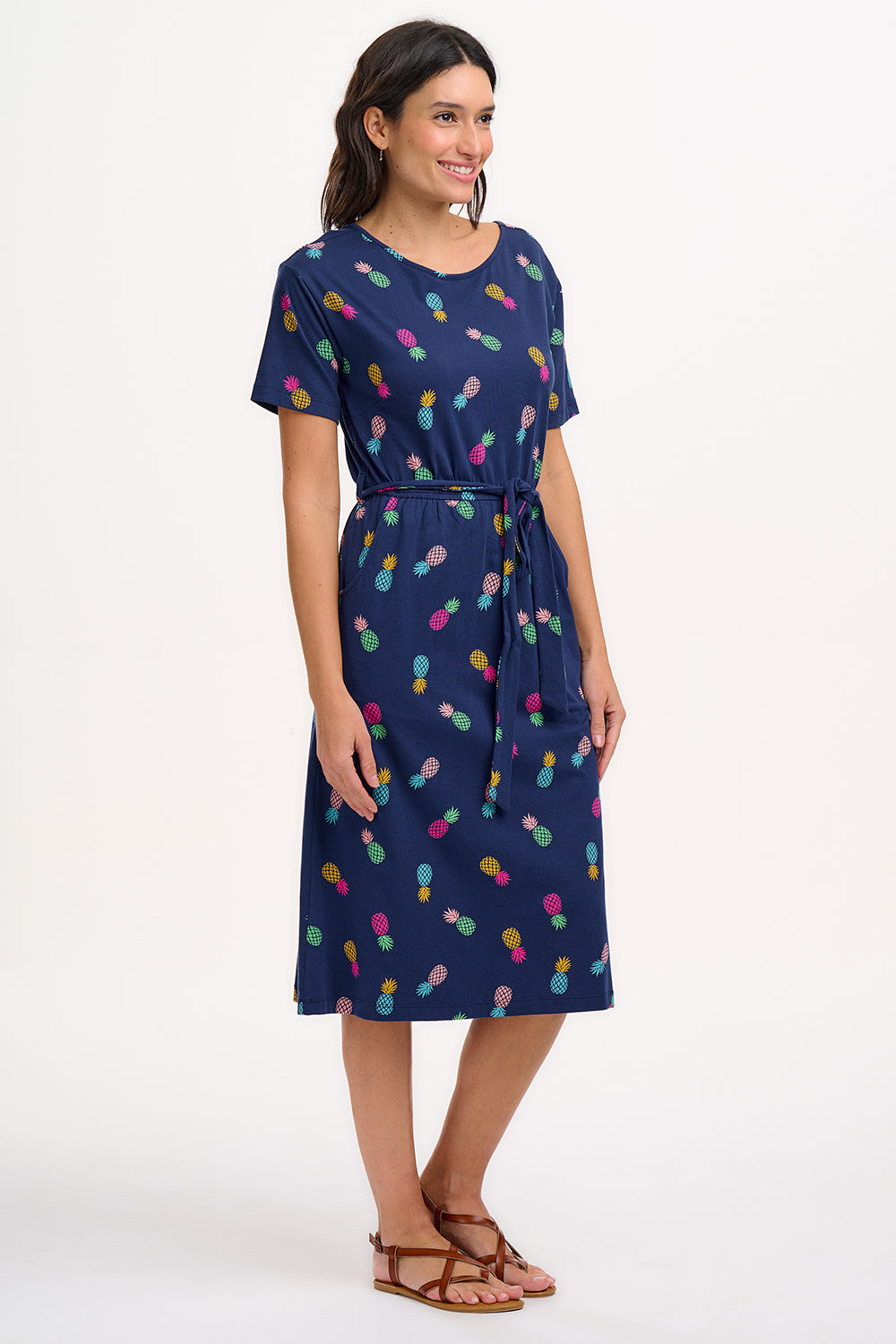 A navy midi jersey dress with a multicolor pineapple print, featuring a low scoop back, short sleeves, and an elasticated waistband with a tie belt. The dress is modeled by a woman standing against a white background.