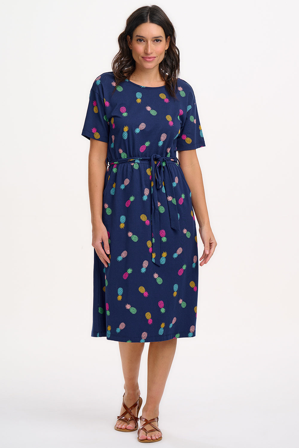 A navy midi jersey dress with a multicolor pineapple print, featuring a low scoop back, short sleeves, and an elasticated waistband with a tie belt. The dress is modeled by a woman standing against a white background.