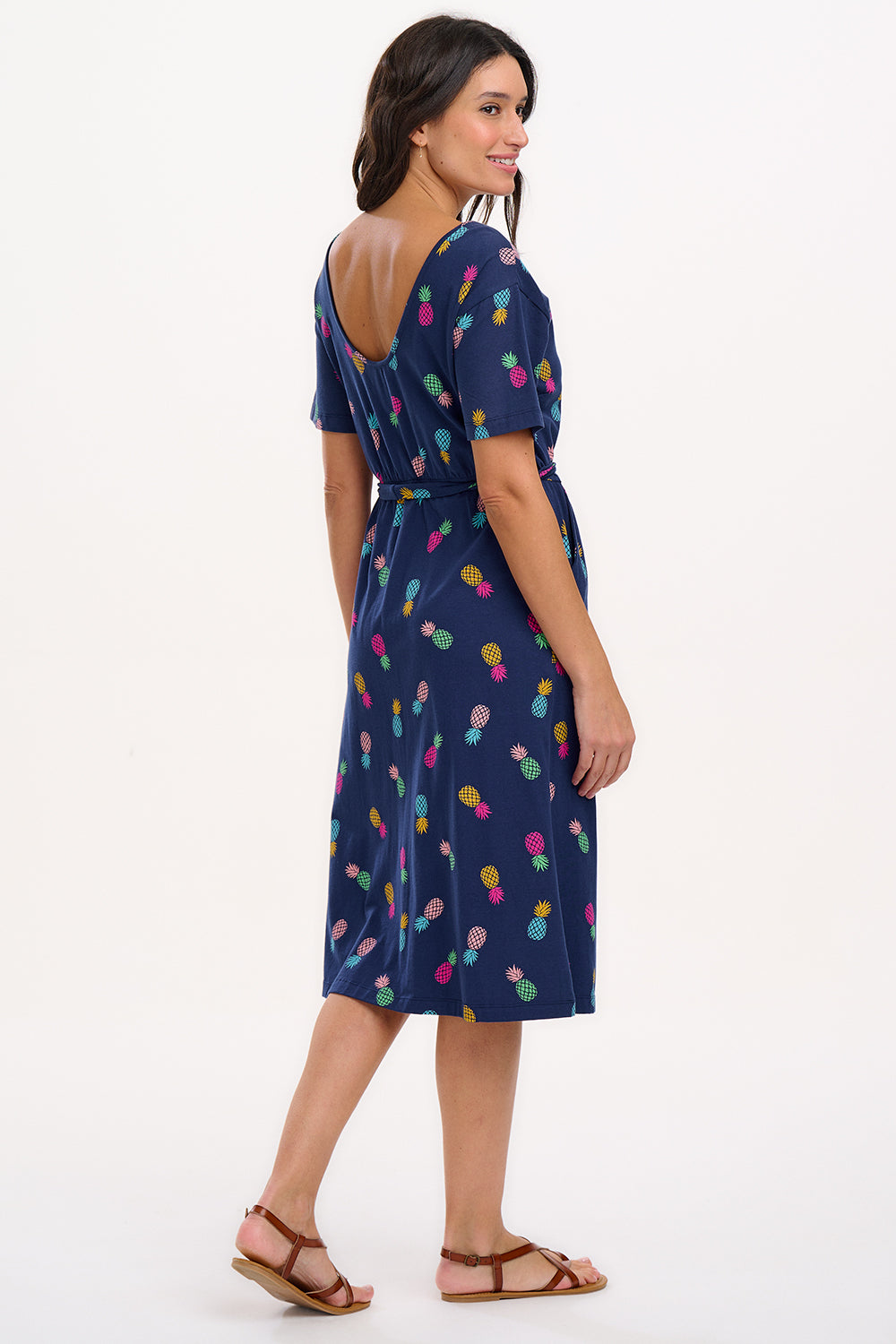 A navy midi jersey dress with a multicolor pineapple print, featuring a low scoop back, short sleeves, and an elasticated waistband with a tie belt. The dress is modeled by a woman standing against a white background.