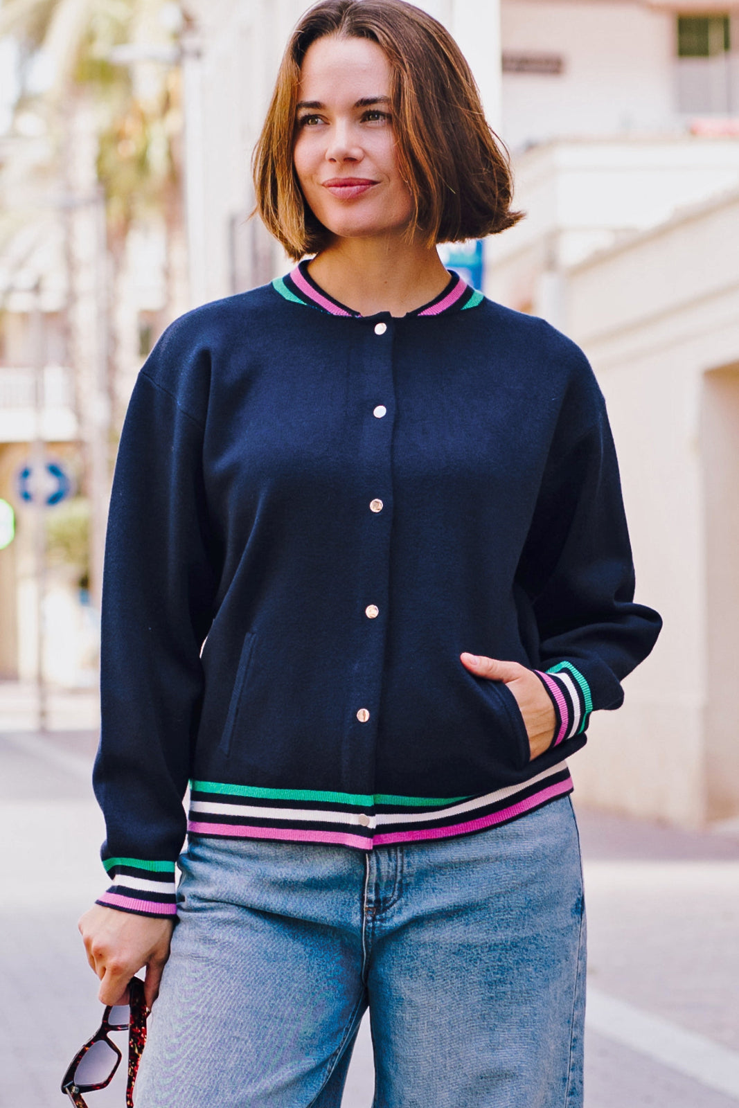 A woman wearing a navy knitted bomber jacket with colorful stripes around the neck, hem, and sleeve cuffs, paired with blue jeans.