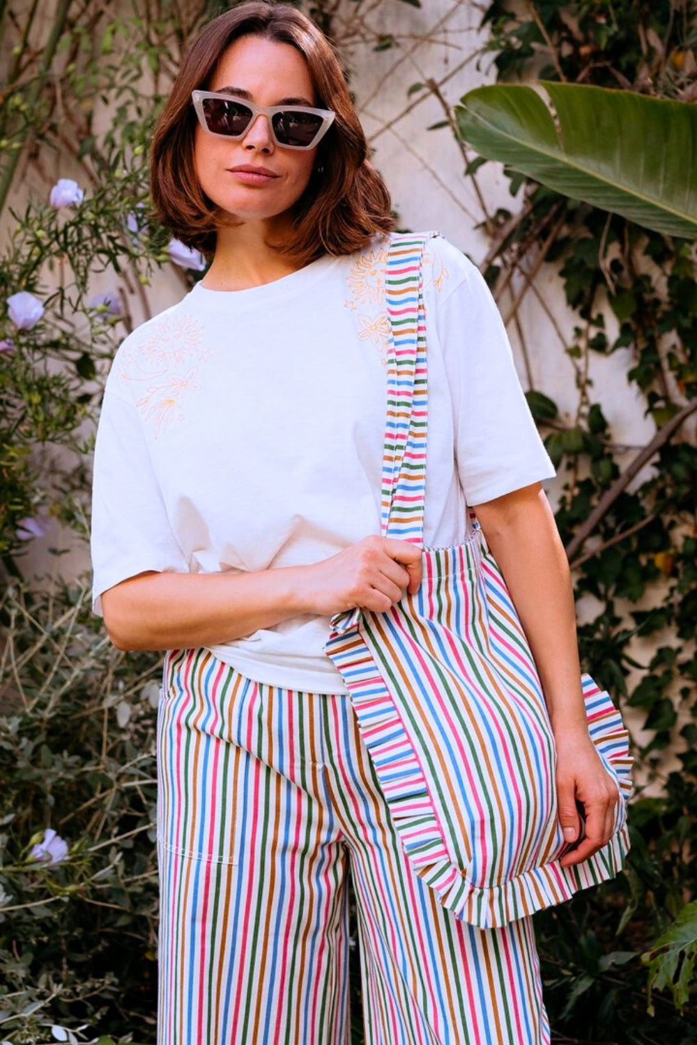 Woman wearing a white t-shirt and colorful striped pants holding a matching bag outdoors.