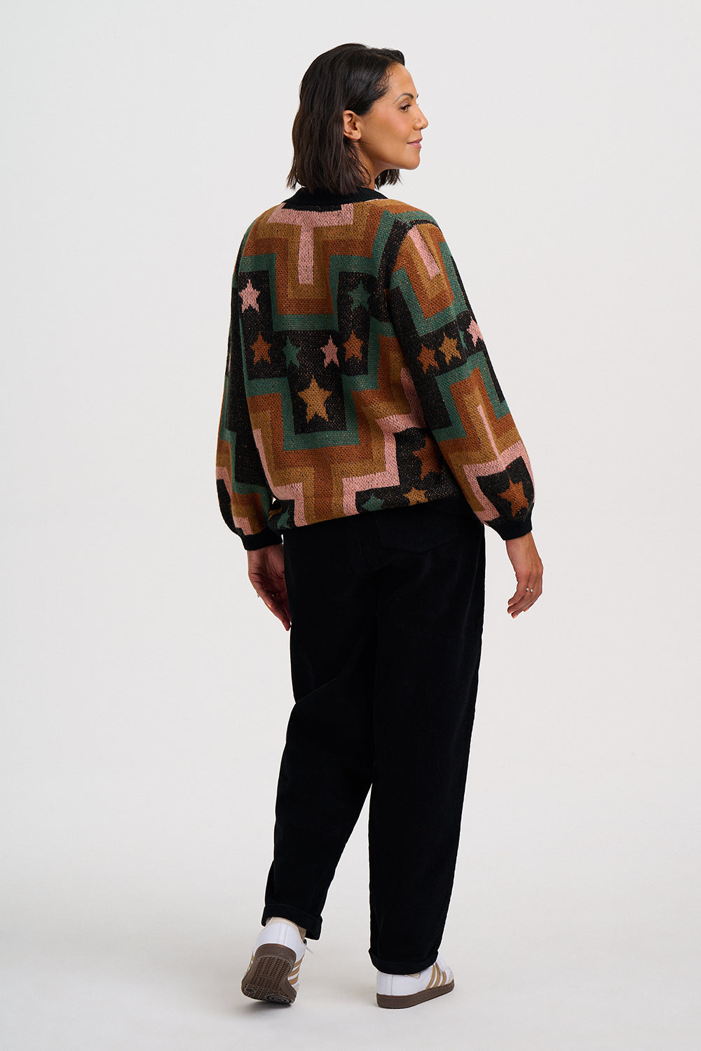 A woman is shown from the back wearing a long-sleeve knit sweater with a geometric pattern in brown, green, pink, and black, paired with black trousers and brown shoes, against a plain white studio background.