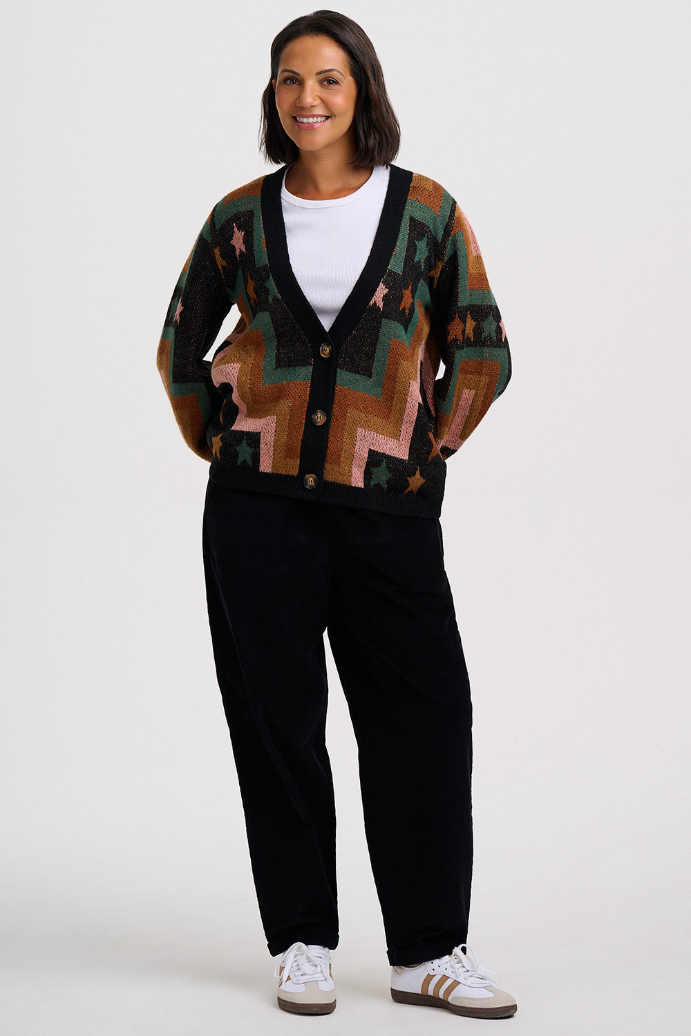 A woman modeling a long-sleeve knit cardigan with a geometric zigzag pattern in brown, green, pink, and black, worn over a white t-shirt and black pants.