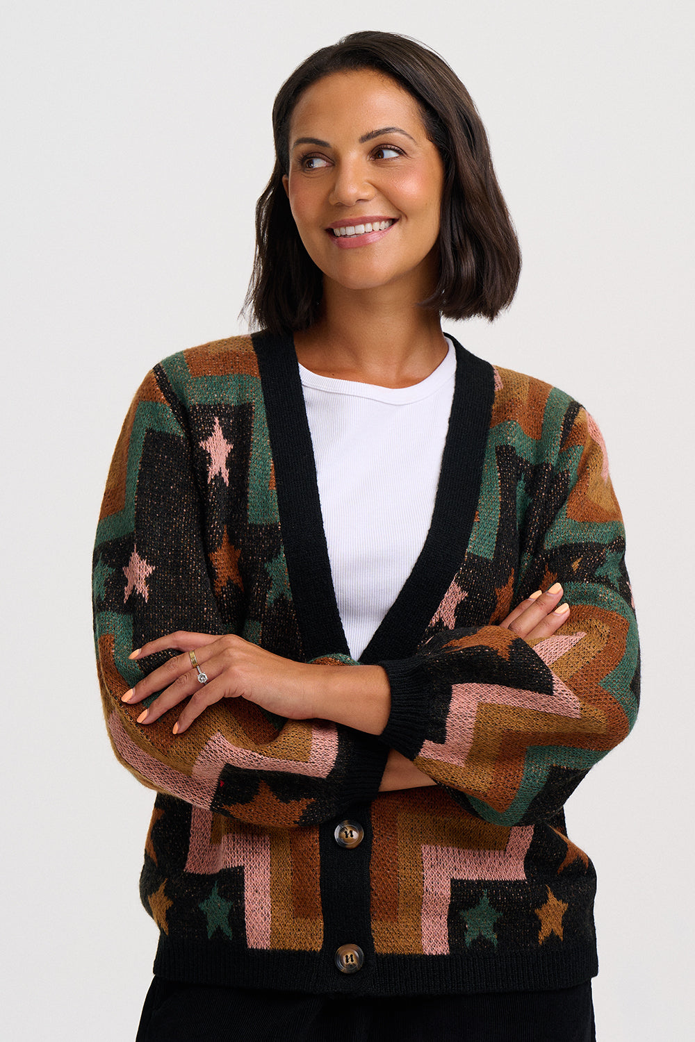 A woman wearing a multicolor knit cardigan with geometric patterns and stars, featuring black trim and a white t-shirt underneath, against a plain light gray background.