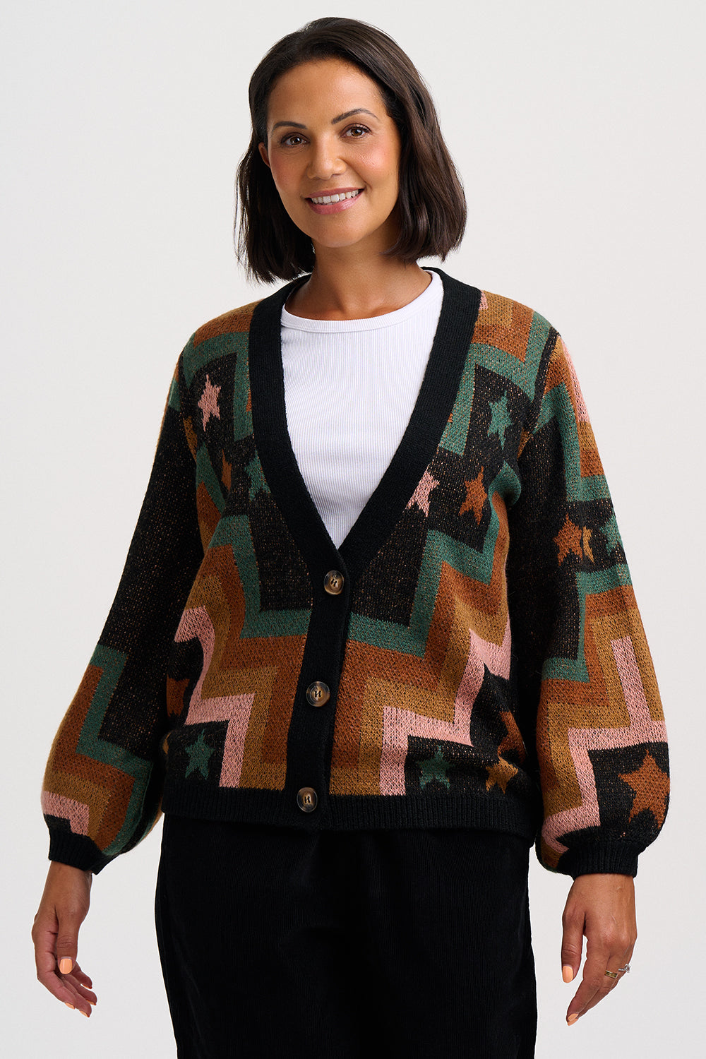 A woman wearing a long-sleeve knit cardigan with a geometric zigzag and star pattern in brown, green, pink, and black, layered over a white top and black pants, against a light gray studio background.