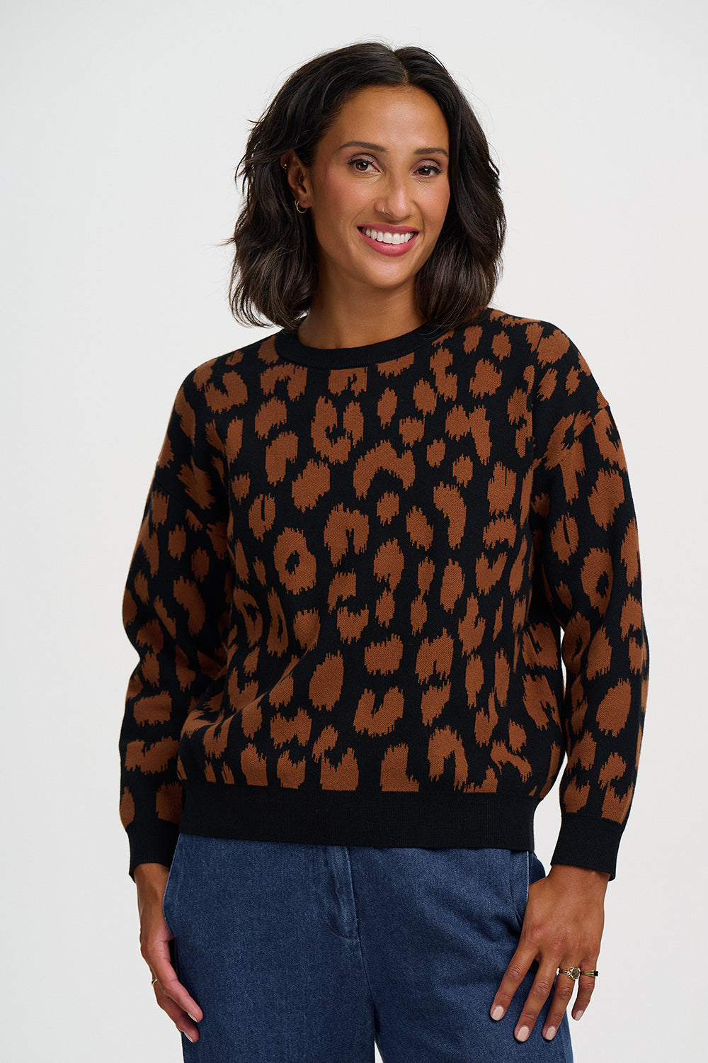 A woman wearing a black crewneck sweater with a brown ikat-style leopard pattern, paired with blue jeans, photographed against a light gray studio background.