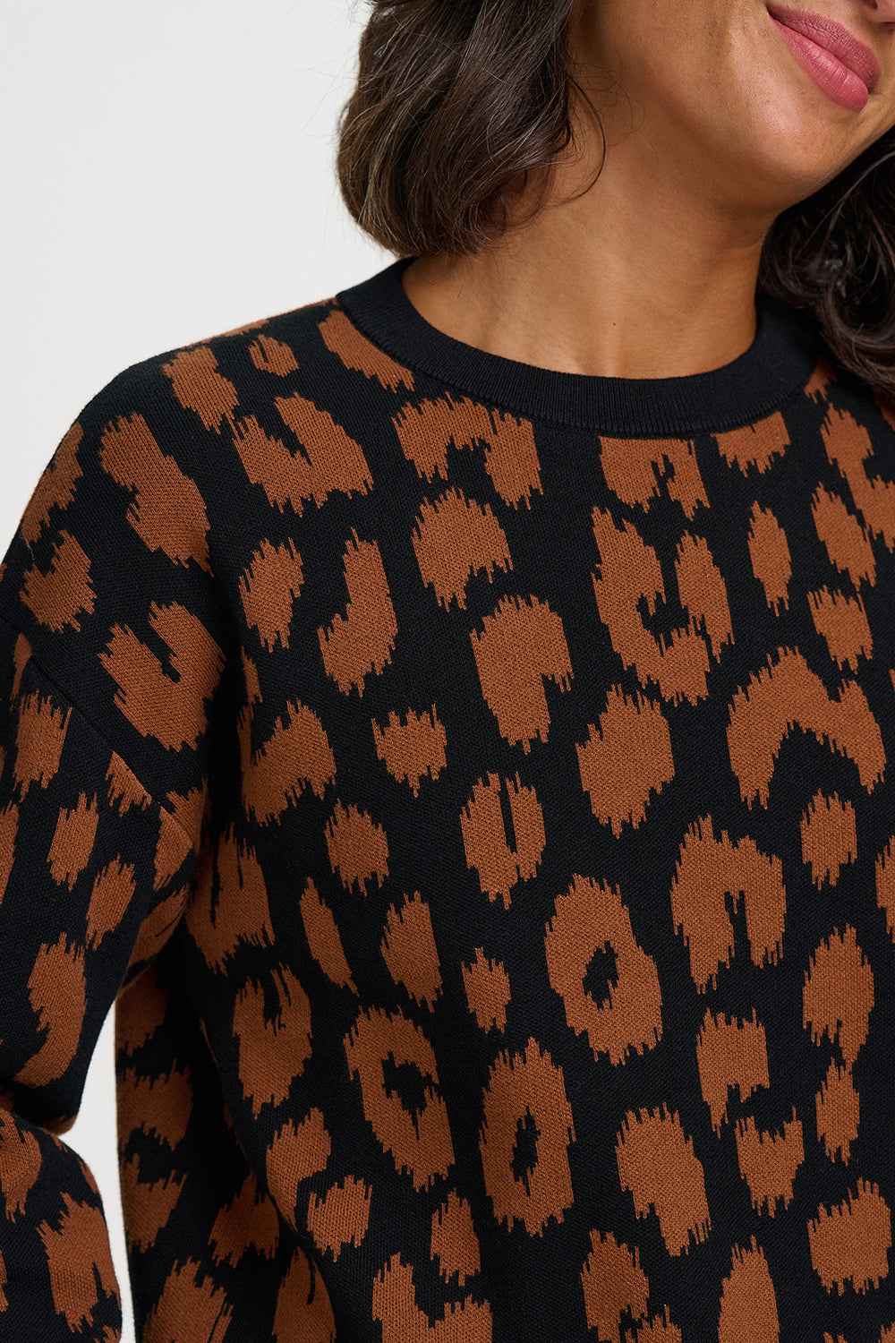 A close-up of a woman wearing a black crewneck sweater with a brown abstract leopard-like pattern, cropped from the mouth down against a plain light background.