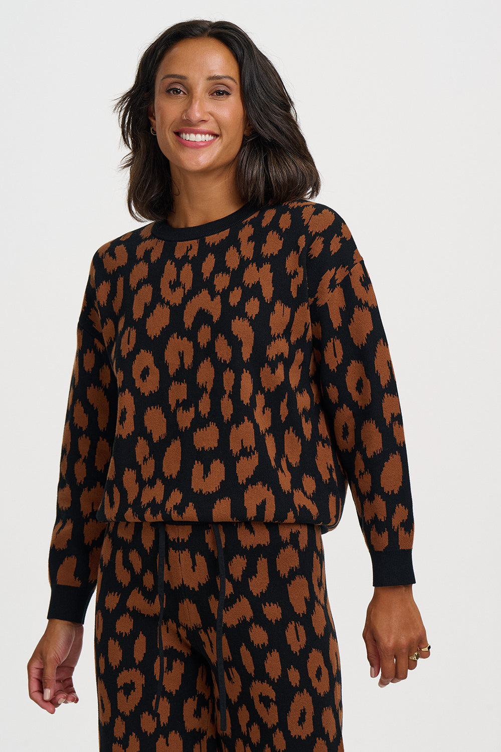 A woman modeling a matching two-piece lounge set featuring a black base with a brown leopard print pattern, including a long-sleeve top and pants, against a plain light gray background.
